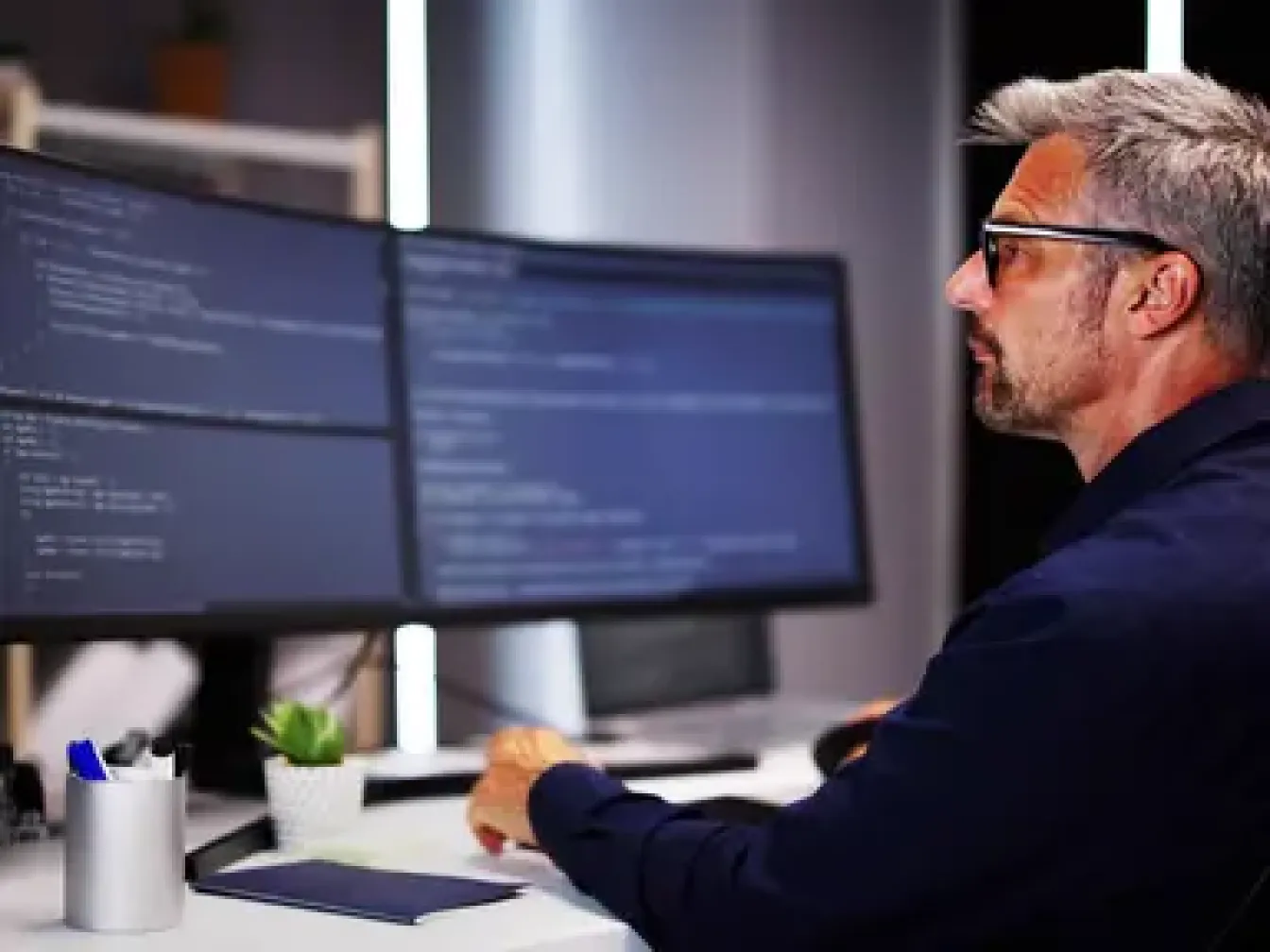 A man with white hair and glasses in front of two monitors showing some code