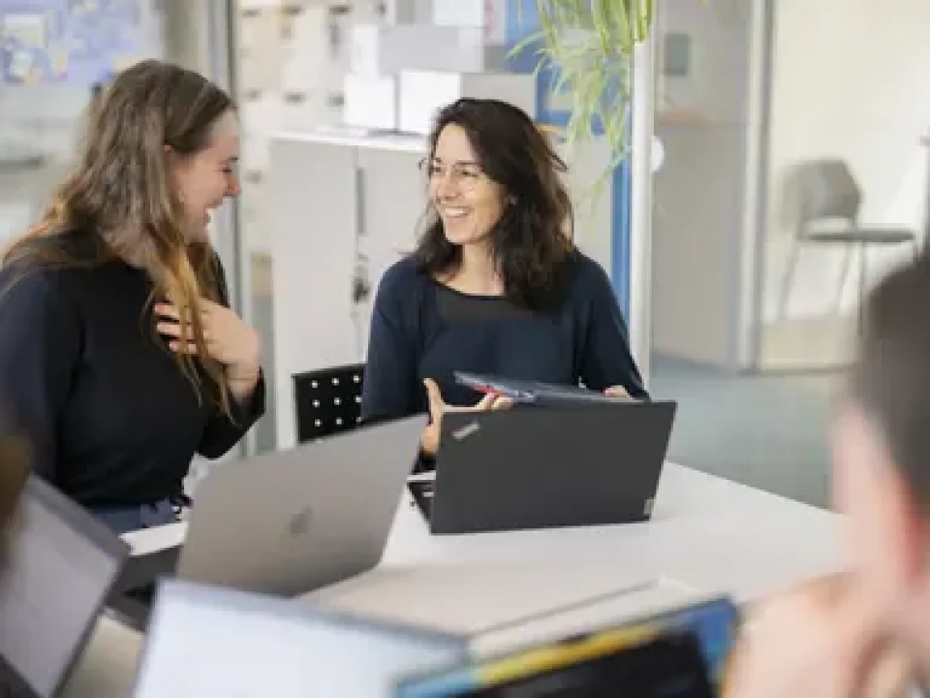 Two women each one in front of a laptop, they are looking to each other and smiling