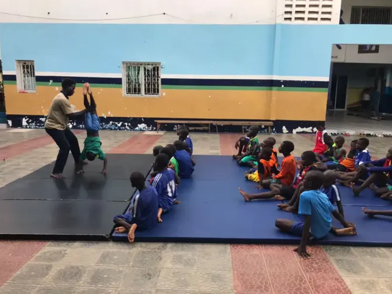 A coach working gymnastic mouvement with a kid in front of other kids sitting in a gym